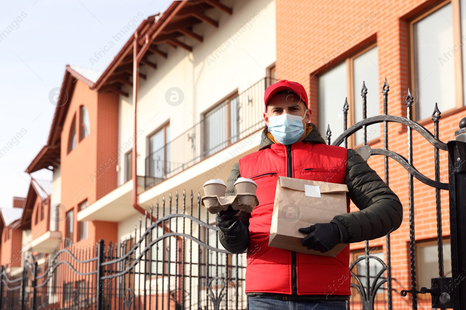 Photo of Courier in medical mask holding takeaway food and drinks near house outdoors. Delivery service during quarantine due to Covid-19 outbreak