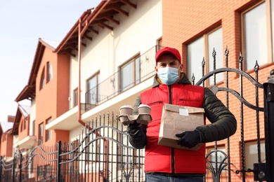 Photo of Courier in medical mask holding takeaway food and drinks near house outdoors. Delivery service during quarantine due to Covid-19 outbreak