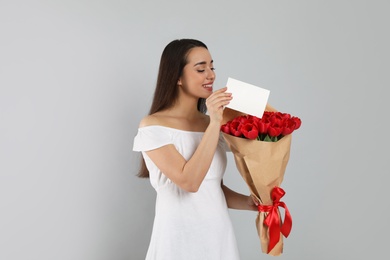 Happy woman with red tulip bouquet and greeting card on light grey background. 8th of March celebration