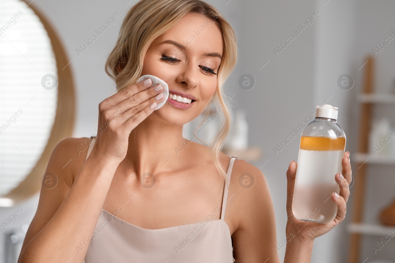 Photo of Smiling woman removing makeup with cotton pad and holding bottle indoors