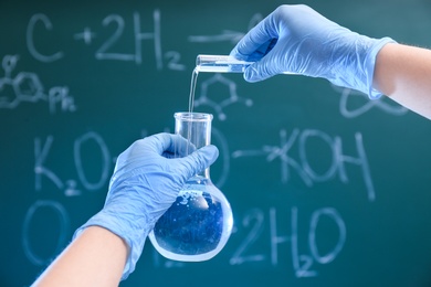 Scientist pouring liquid into flask against chalkboard, closeup. Chemistry glassware