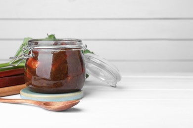 Photo of Jar of tasty rhubarb jam, stems and spoon on white wooden table. Space for text