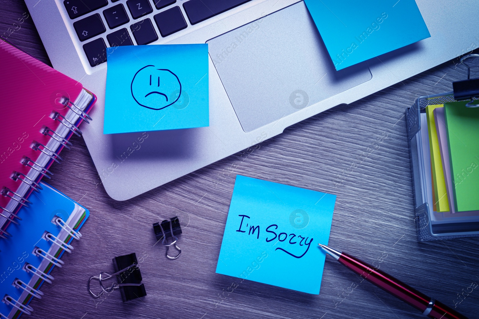 Image of Sticky notes with word Sorry and drawn sad face on workplace. Laptop and stationery on wooden table, flat lay