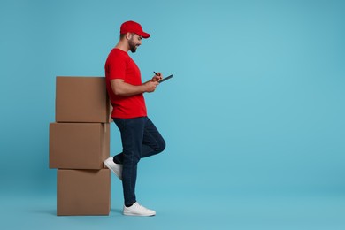 Photo of Happy young courier with clipboard and stack of parcels on light blue background, space for text