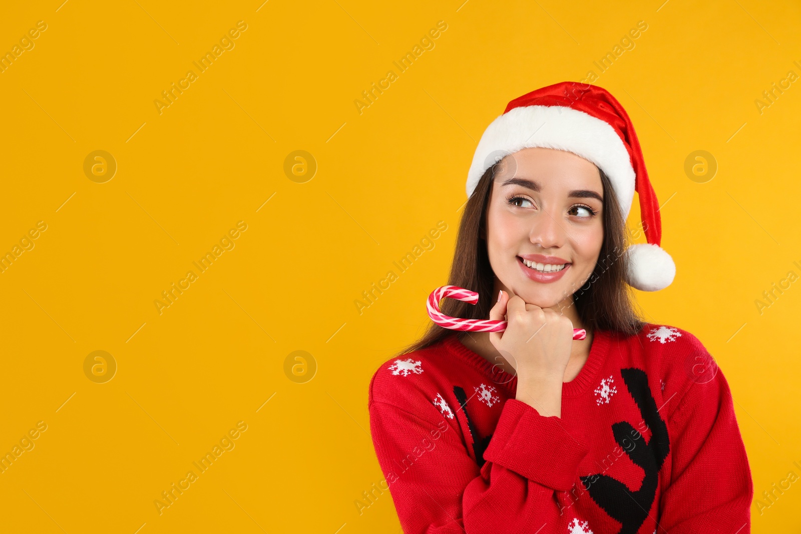 Photo of Young woman in Christmas sweater and Santa hat holding candy cane on yellow background, space for text