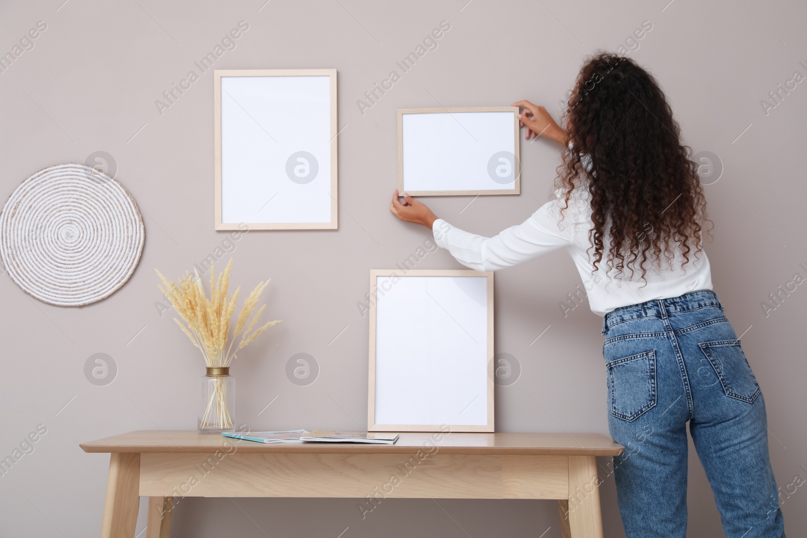 Photo of African American woman hanging empty frame on pale rose wall over table in room, back view. Mockup for design