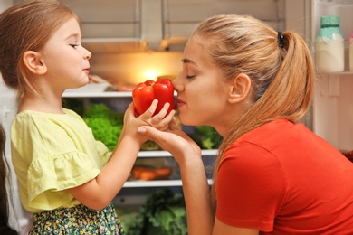Photo of Young mother and daughter with fresh bell pepper near refrigerator at home