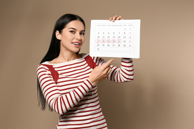 Young woman holding calendar with marked menstrual cycle days on beige background