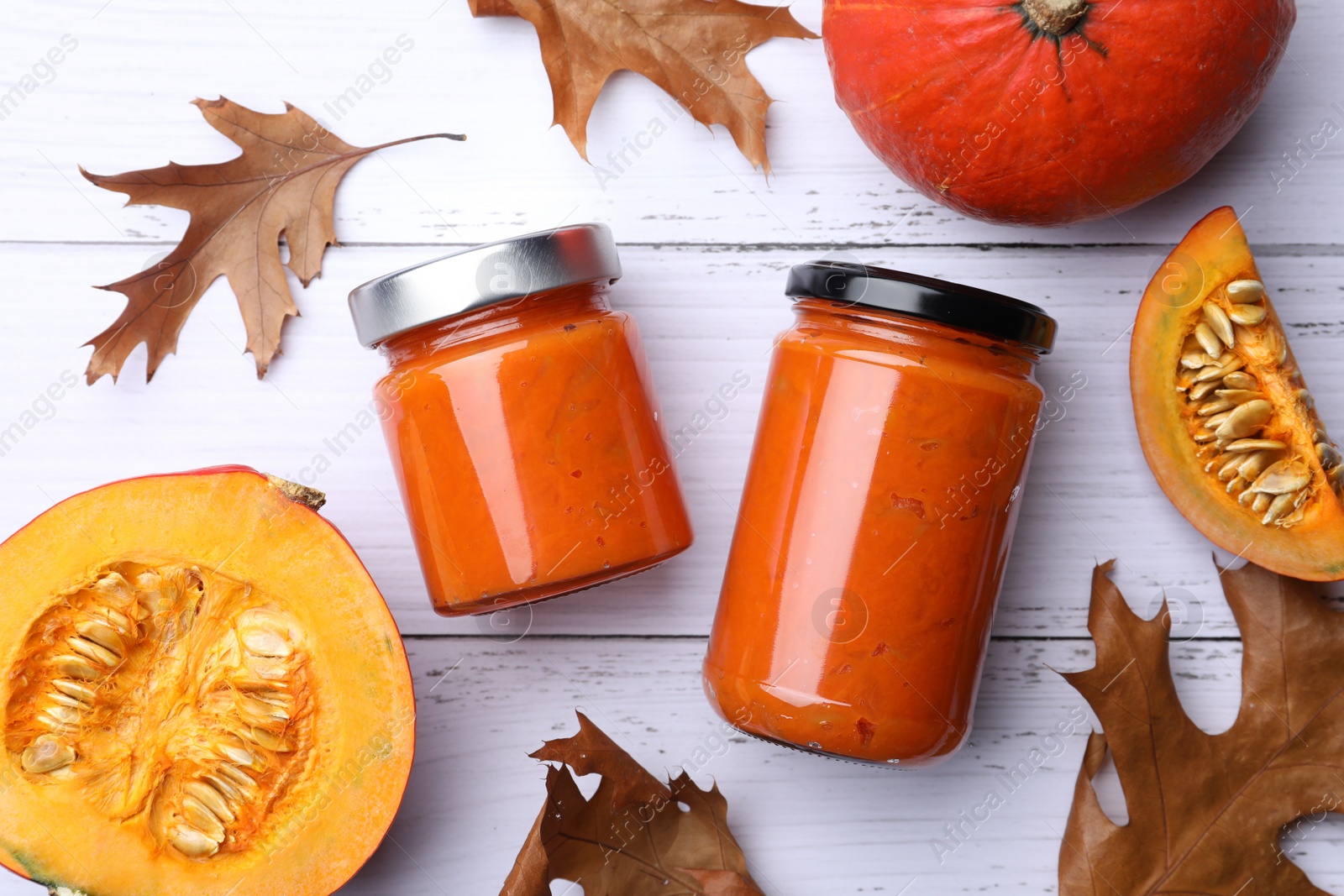 Photo of Jars of delicious pumpkin jam, fresh pumpkin and dry leaves on white wooden table, flat lay