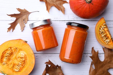 Jars of delicious pumpkin jam, fresh pumpkin and dry leaves on white wooden table, flat lay