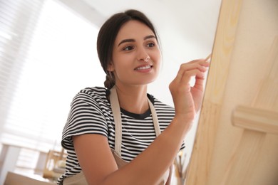 Photo of Young woman drawing on canvas with brush indoors