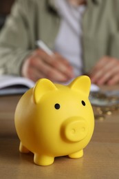 Photo of Man at wooden table, focus on yellow piggy bank