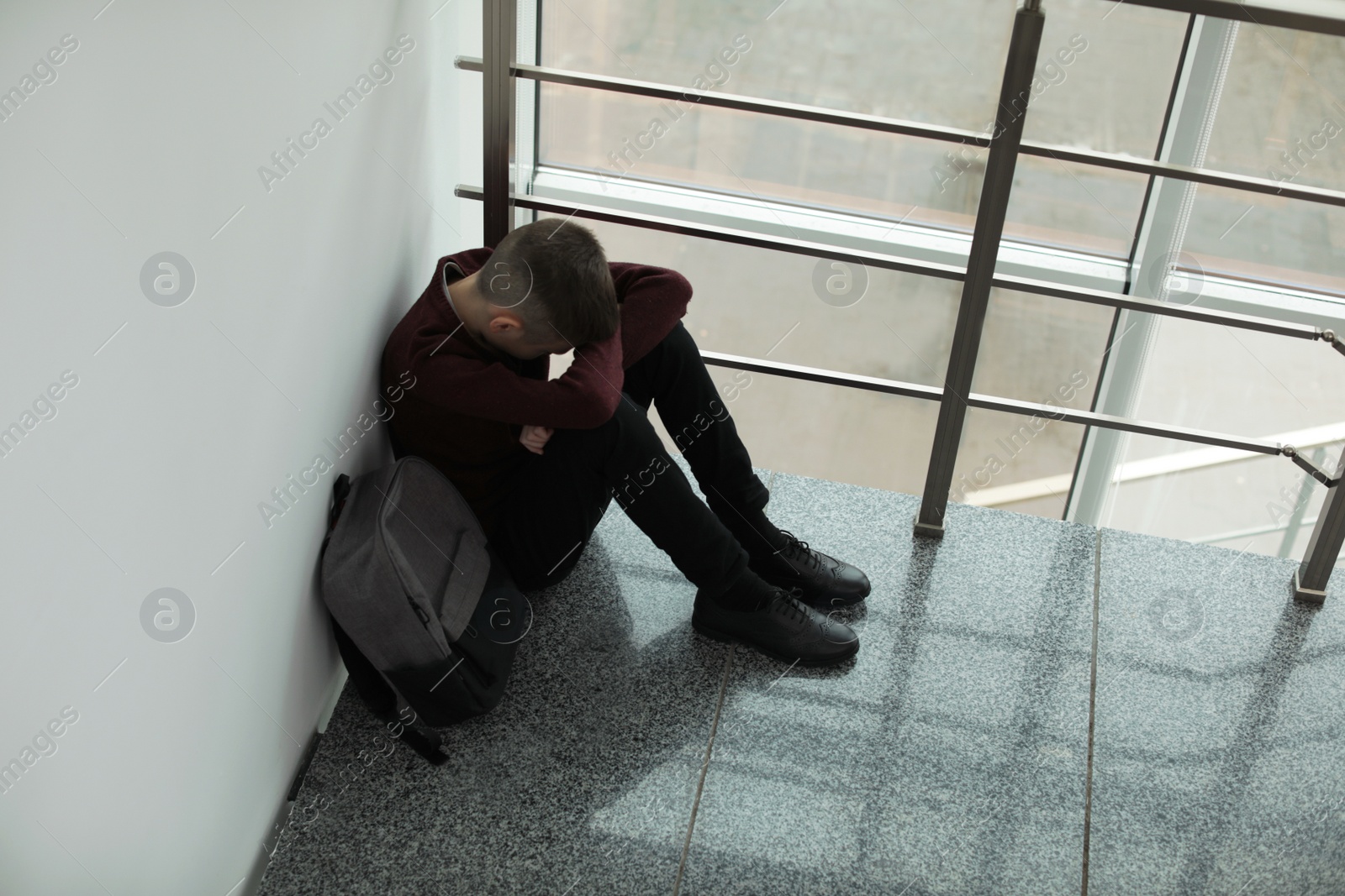 Photo of Upset boy with backpack sitting near window indoors. Space for text