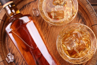 Whiskey with ice cubes in glasses and bottle on wooden barrel, flat lay