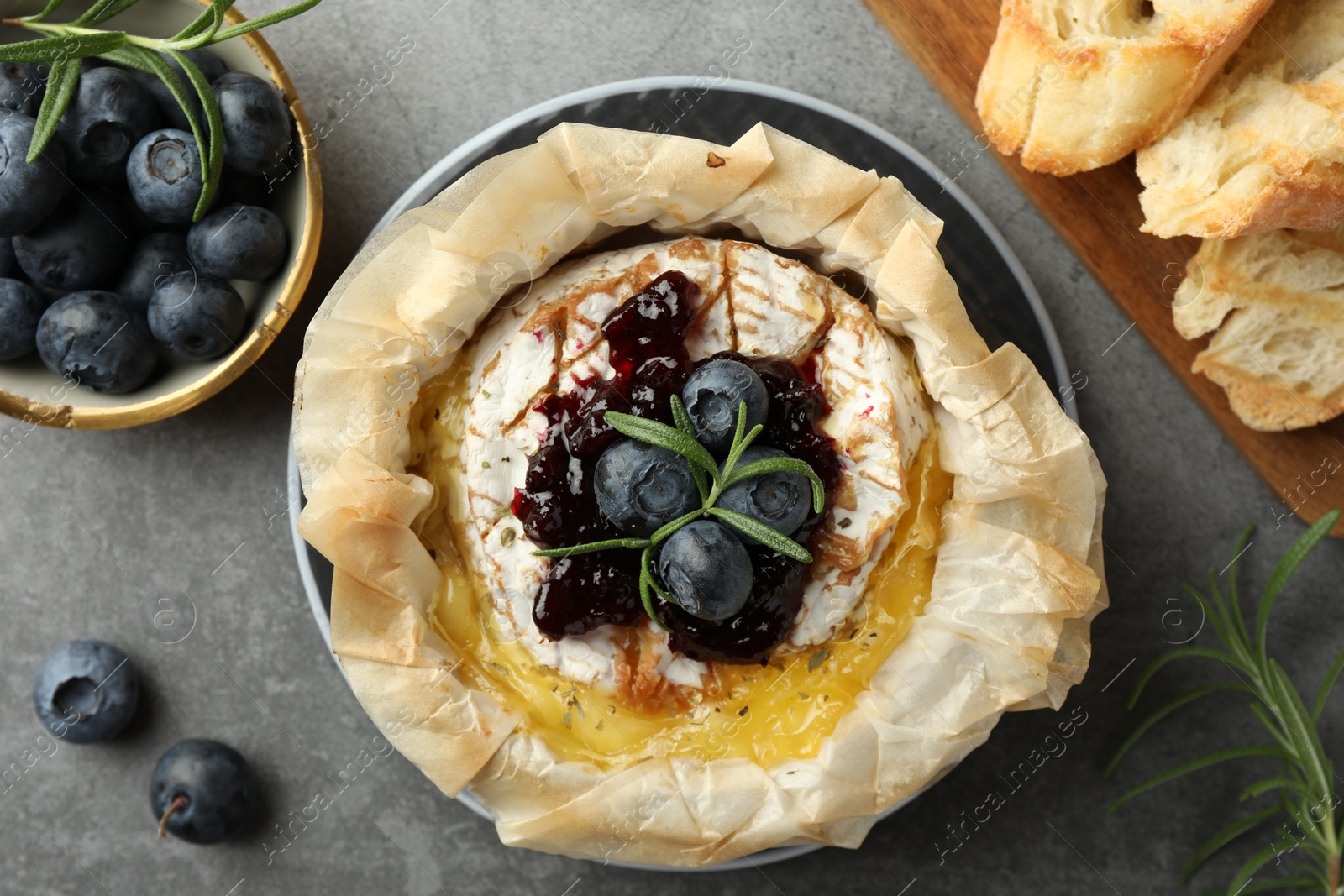 Photo of Tasty baked brie cheese with blueberries, jam and rosemary on grey table, flat lay