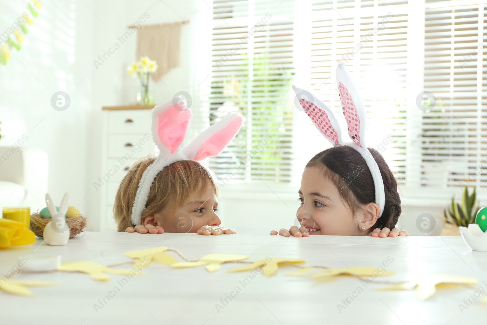 Photo of Cute children wearing bunny ears headbands at table with Easter eggs, indoors