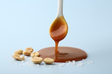 Spoon with tasty salted caramel, peanuts and salt on light blue background, closeup