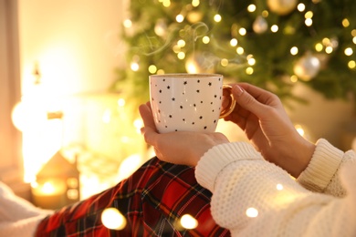Photo of Woman with cup of hot drink near Christmas tree indoors, closeup