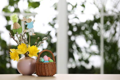 Festive composition with Easter eggs in wicker basket on table against blurred window, space for text