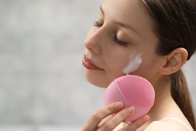 Young woman washing face with brush and cleansing foam on light background, closeup. Space for text