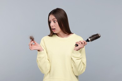 Emotional woman holding brush with lost hair on light grey background. Alopecia problem