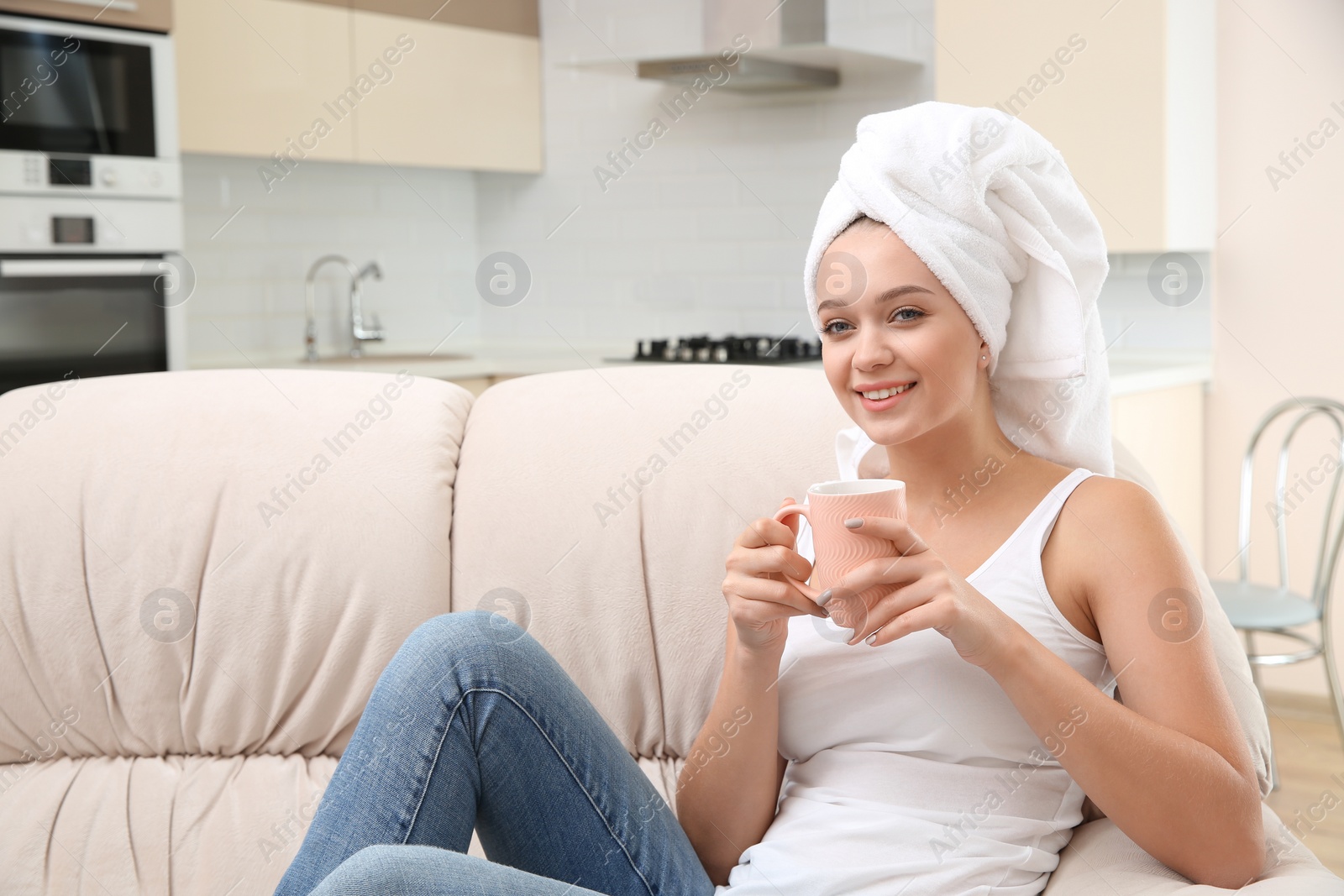 Photo of Beautiful woman with hair wrapped in towel sitting on sofa and drinking tea at home