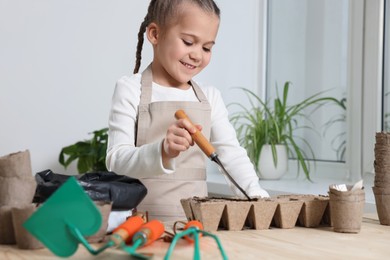 Photo of Little girl adding soil into peat pots at wooden table indoors. Growing vegetable seeds