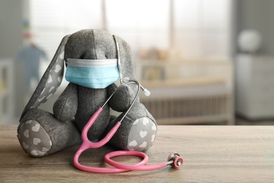 Image of Toy bunny with protective mask and stethoscope on wooden table indoors, space for text. Pediatrician practice