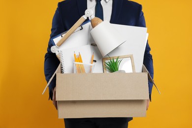 Photo of Unemployed man with box of personal office belongings on orange background, closeup