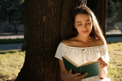 Beautiful young woman reading book near tree in park