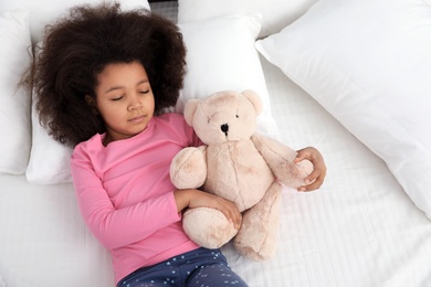 Photo of Cute little African-American girl with teddy bear sleeping in bed, top view