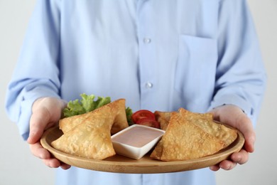 Photo of Woman holding plate with delicious samosas and sauce on light grey background, closeup