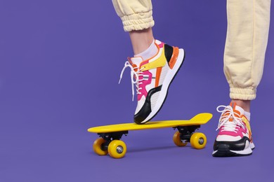 Photo of Woman in new stylish sneakers standing on skateboard against purple background, closeup. Space for text