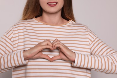 Young woman making heart with hands on light grey background, closeup. Volunteer concept