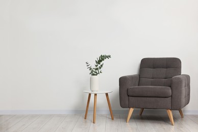 Photo of Comfortable armchair, side table and eucalyptus near white wall indoors, space for text