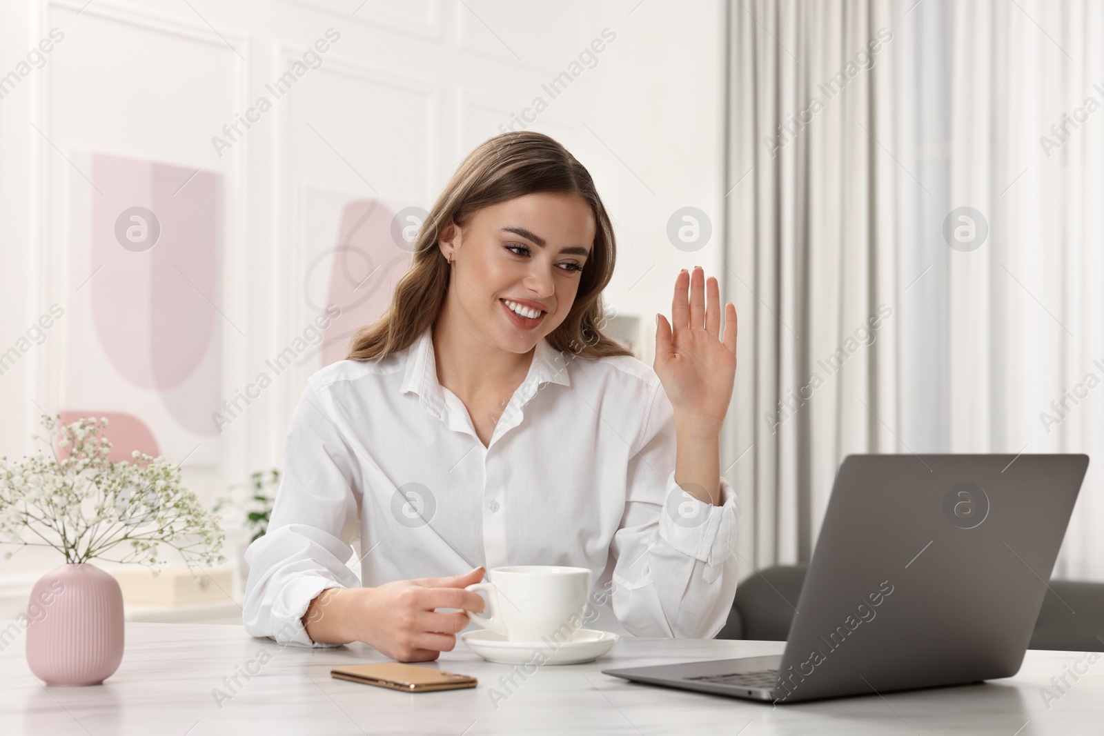 Photo of Happy woman with cup of drink using laptop at white table
