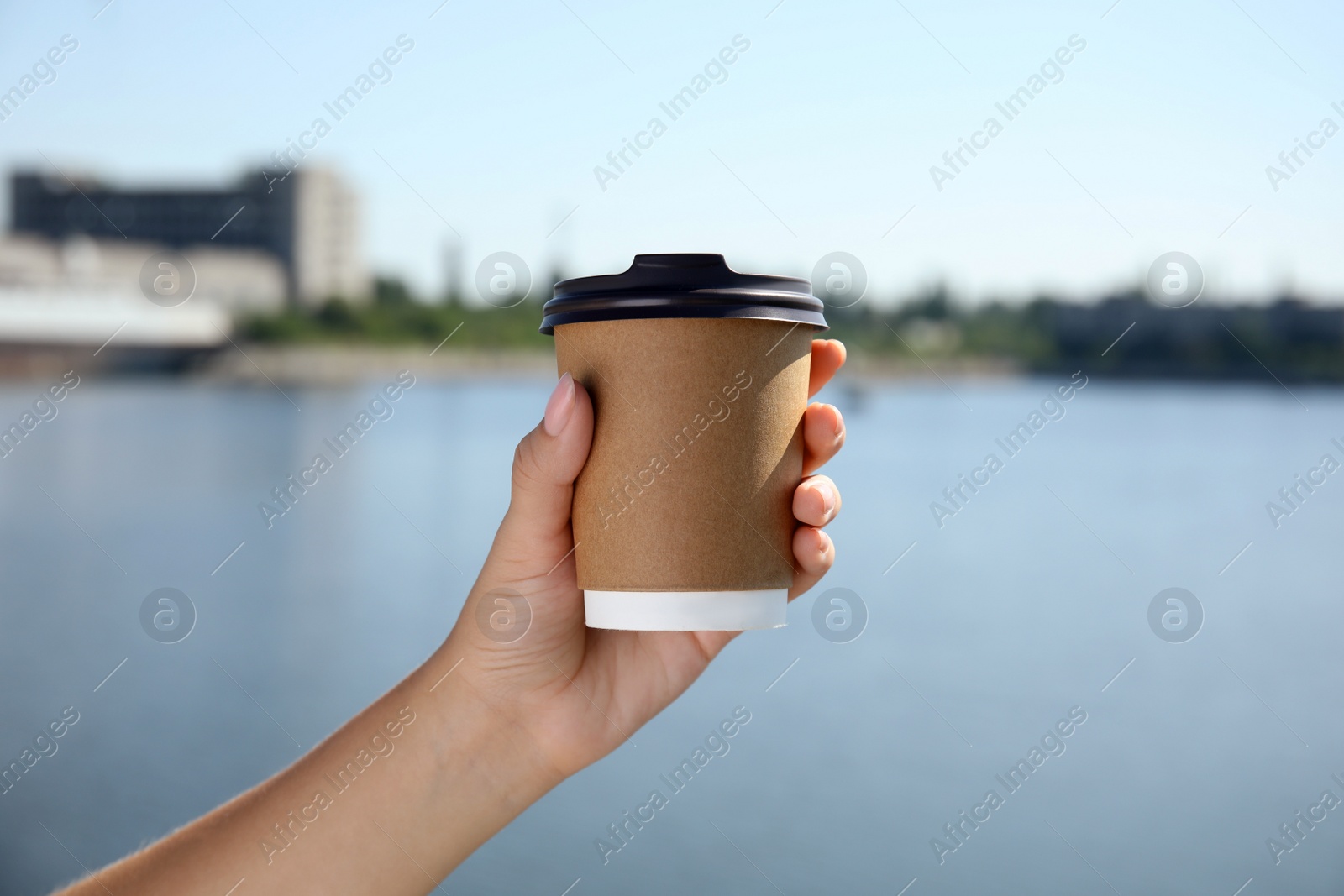 Photo of Woman holding takeaway cardboard coffee cup with plastic lid against blue river, closeup