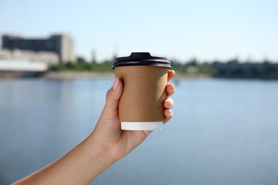 Woman holding takeaway cardboard coffee cup with plastic lid against blue river, closeup