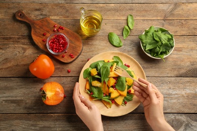 Photo of Woman eating delicious persimmon salad with pomegranate and spinach at wooden table, top view
