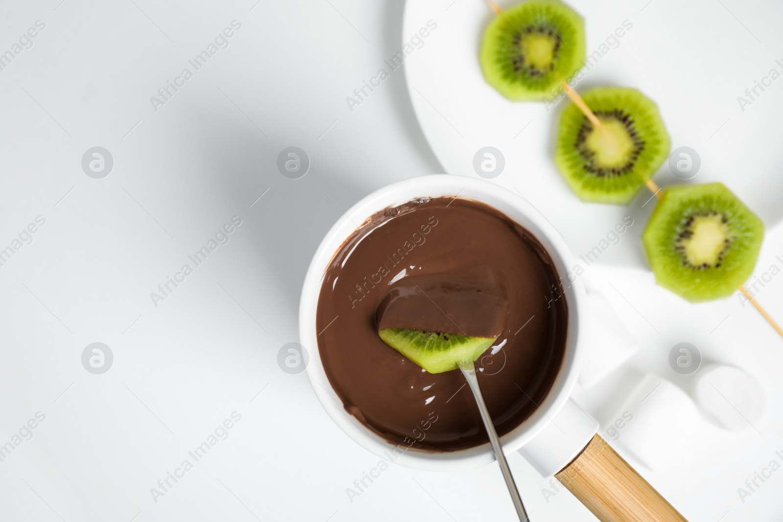 Photo of Dipping kiwi into fondue pot with chocolate on white table, flat lay. Space for text