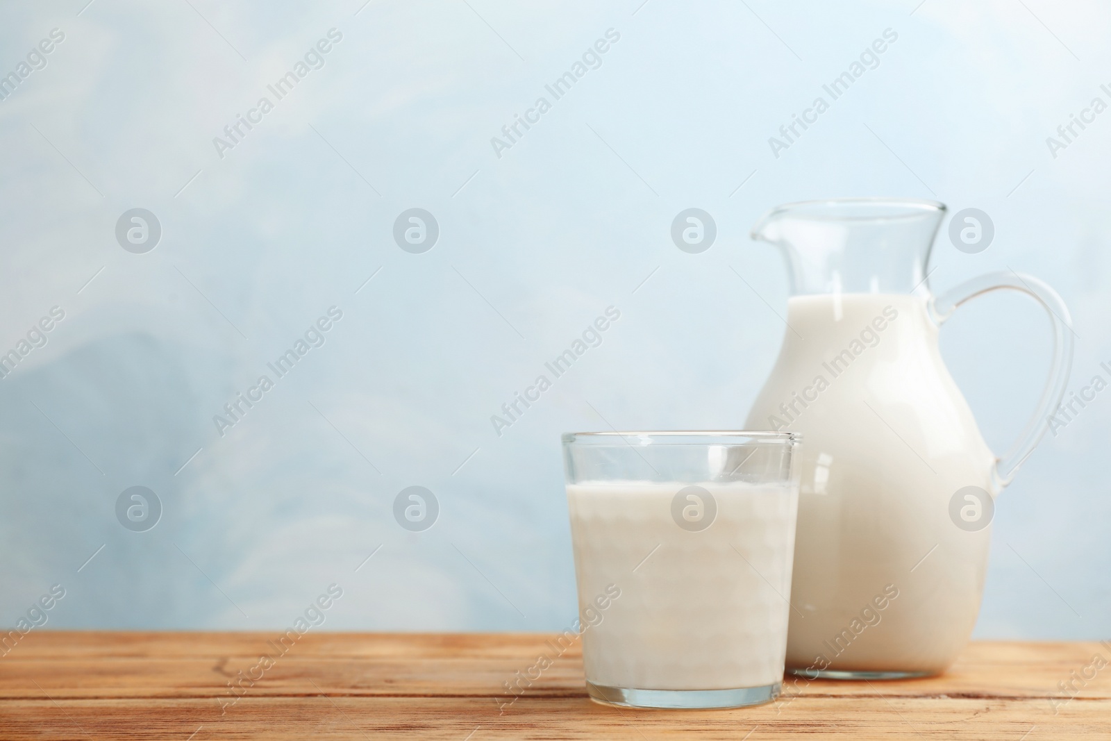 Photo of Glass and jug of fresh milk on wooden table against color background. Space for text