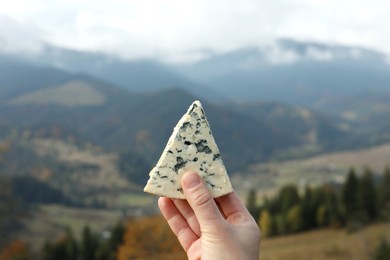 Woman holding piece of delicious cheese against mountain landscape, closeup
