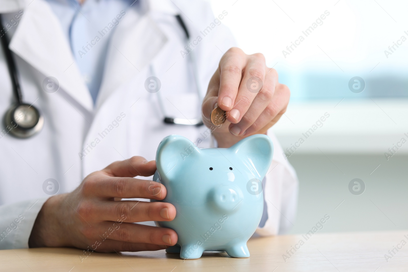 Photo of Doctor putting coin into piggy bank at wooden table, closeup