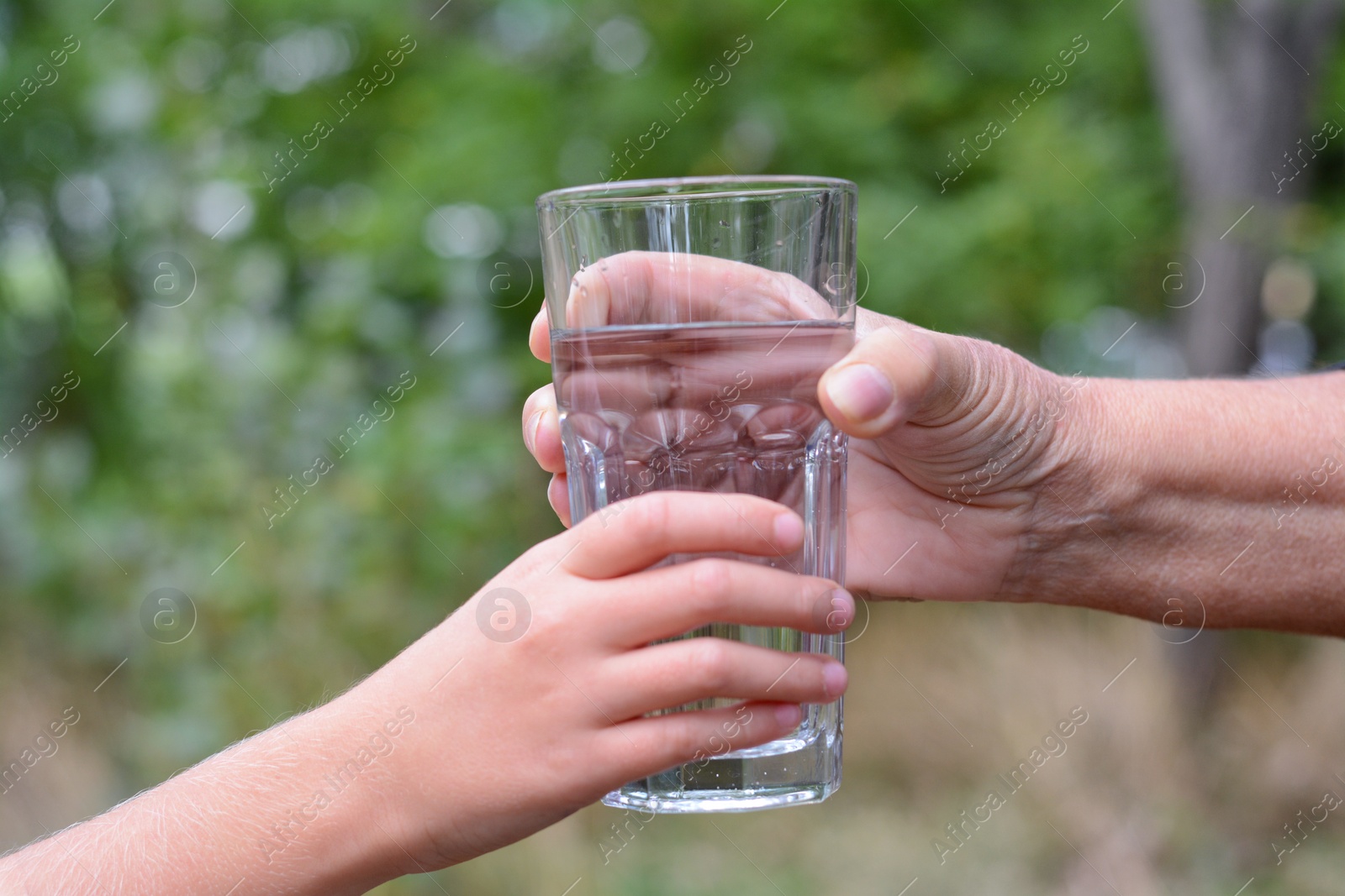 Photo of Child giving glass of water to elderly woman outdoors, closeup