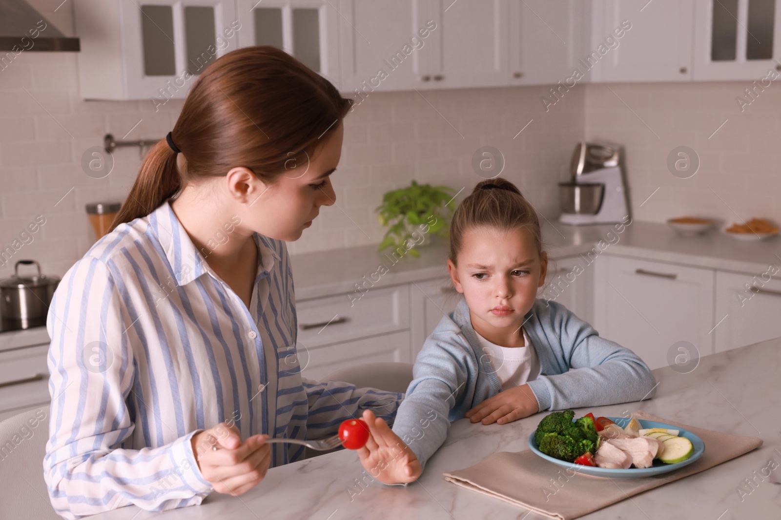 Photo of Mother feeding her daughter in kitchen. Little girl refusing to eat dinner