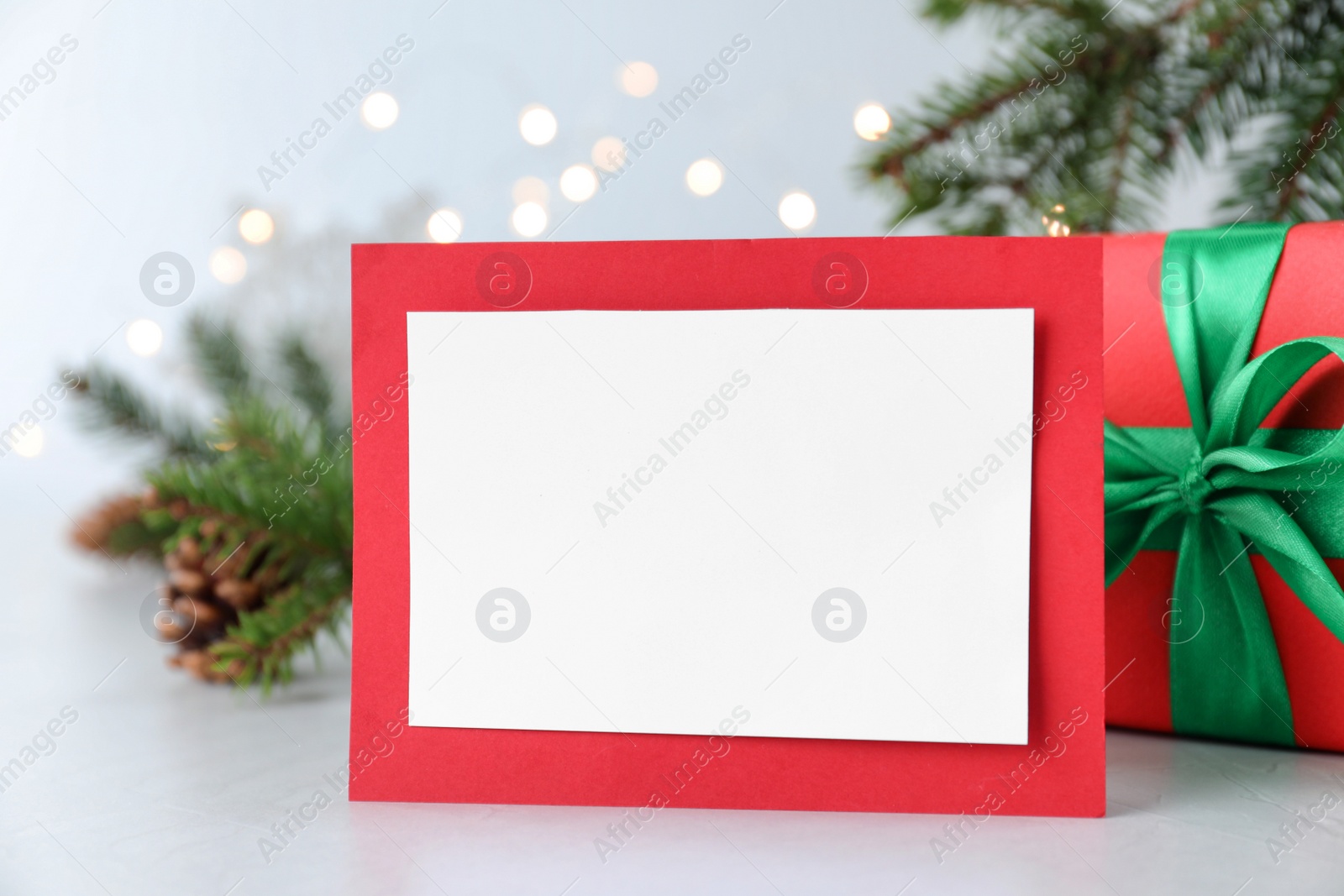 Photo of Blank Christmas card and gift box on table against blurred fairy lights, closeup. Space for text