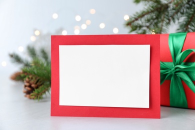 Blank Christmas card and gift box on table against blurred fairy lights, closeup. Space for text