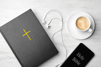 Bible, phone, cup of coffee and earphones on white wooden background, flat lay. Religious audiobook