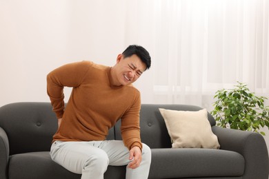 Photo of Asian man suffering from back pain on sofa indoors