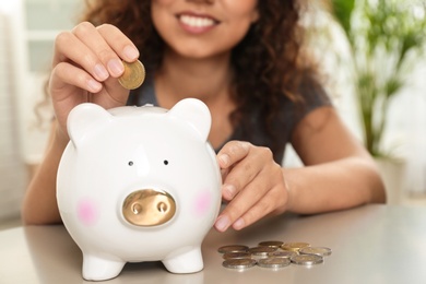 Photo of Happy woman putting coin into piggy bank at table, closeup